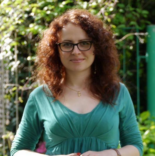 Portret kobiety z rudymi kręconymi włosami, w okularach i zielonej sukience, stojącej na tle zieleni ogrodu.