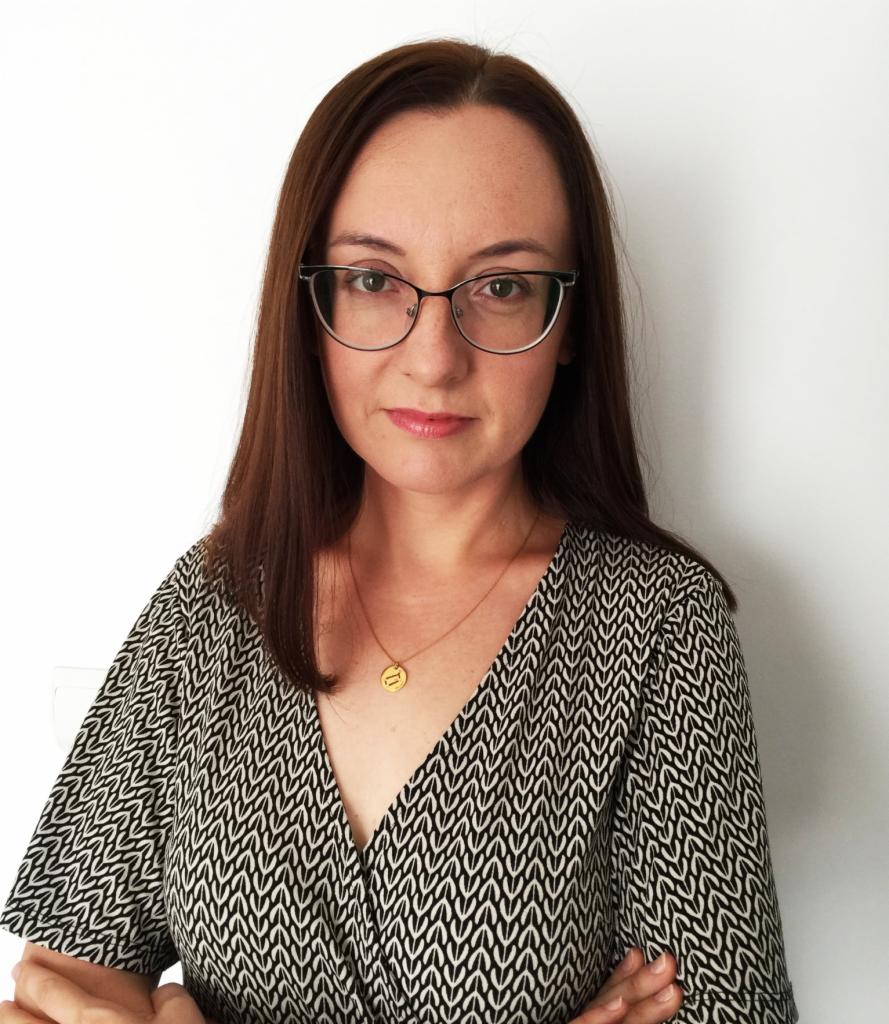 Portret kobiety w okularach, z brązowymi włosami i wzorzystej sukience, na białym tle.