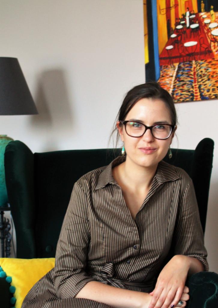 Portret kobiety w okularach siedzącej w zielonym fotelu, z kolorowym obrazem w tle.