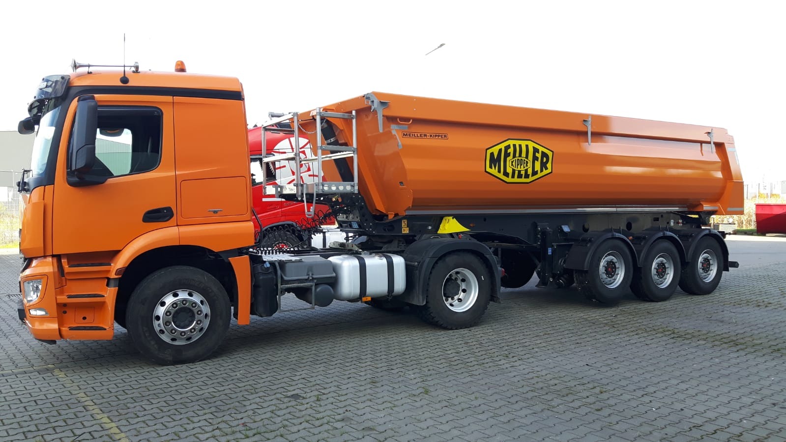 Pomarańczowa ciężarówka wywrotka marki Meller Kipper zaparkowana na szarym bruku, z widocznym logo firmy na burcie, w tle częściowo widoczna czerwona ciężarówka.