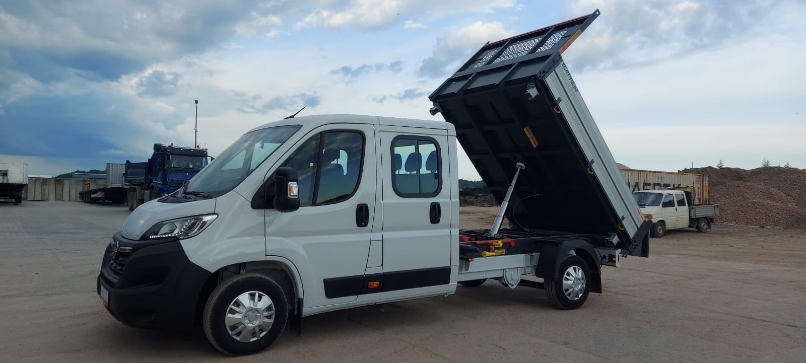 Biały samochód dostawczy z podniesioną skrzynią ładunkową na placu budowy, w tle inne pojazdy i hałda materiału.