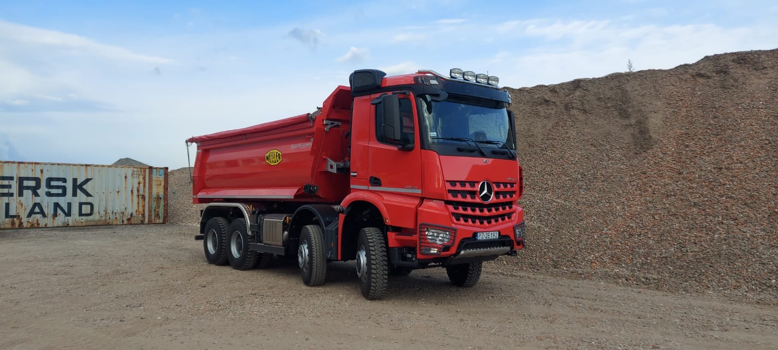 Czerwona wywrotka Mercedes-Benz Arocs na żwirowym podłożu, z kontenerem i hałdą gruzu w tle, pod błękitnym niebem.