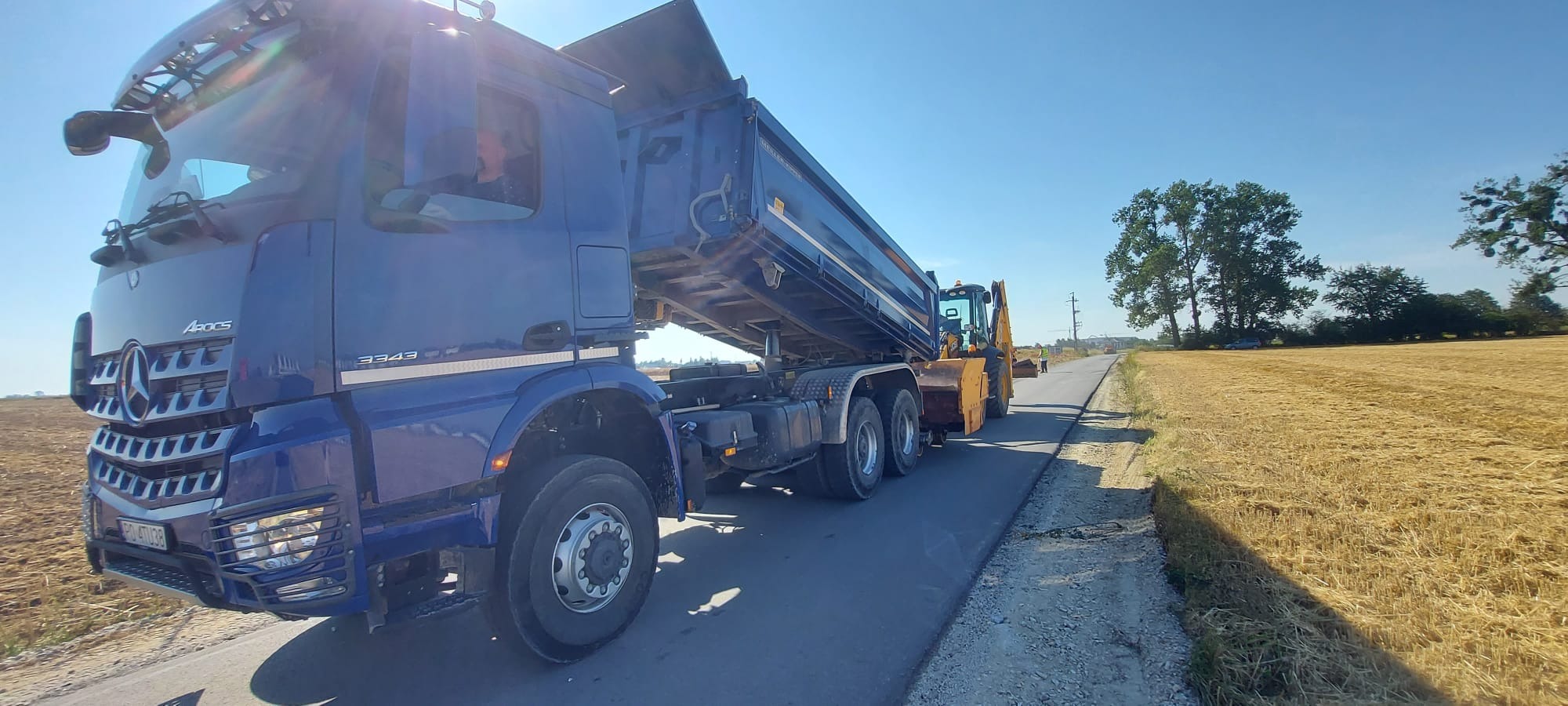 Niebieska ciężarówka wywrotka Mercedes-Benz Arocs 3343 z podniesioną skrzynią ładunkową i żółta koparko-ładowarka na asfalcie, w tle pole i drzewa pod błękitnym niebem.