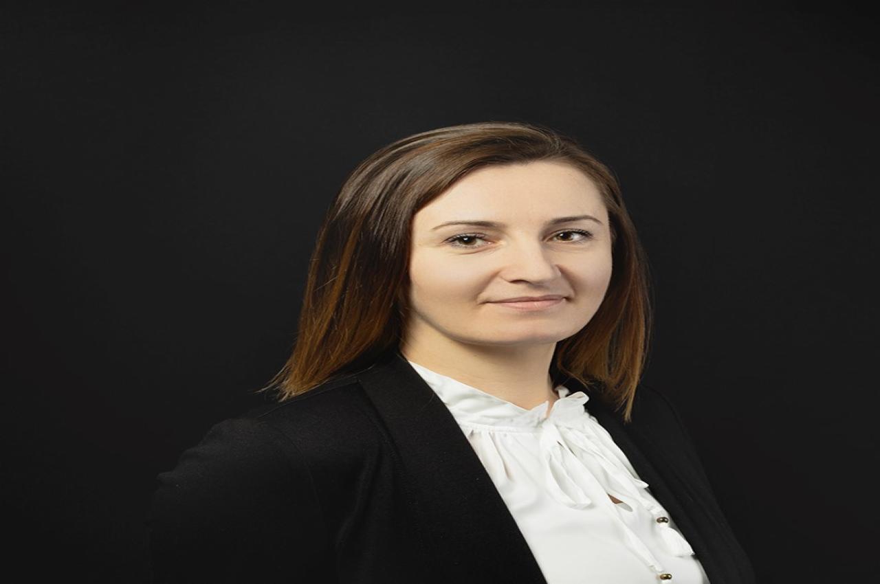 Portret uśmiechniętej kobiety w czarnej marynarce i białej bluzce z wiązaniem pod szyją na ciemnym tle.