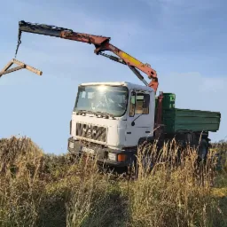 Samochód ciężarowy MAN z dźwigiem HDS podnosi drewnianą konstrukcję w Bytomiu, widok z dołu przez wysokie trawy.