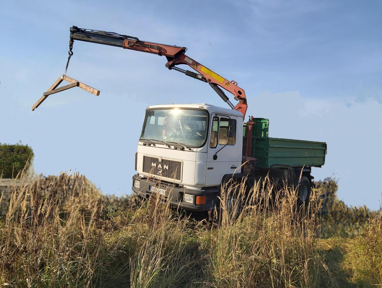 Samochód ciężarowy MAN z dźwigiem HDS podnosi drewnianą konstrukcję w Bytomiu, widok z dołu przez wysokie trawy.