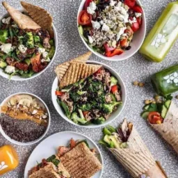 Kompozycja z różnorodnych dań: sałatki z warzywami i serem feta, bowl z bananami i masłem orzechowym, wrap z awokado i warzywami oraz kanapki na tle butelek ze świeżymi sokami.