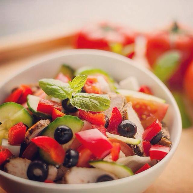 Świeża sałatka z grillowanym kurczakiem, ogórkiem, papryką, pomidorami, oliwkami i serem feta, udekorowana gałązką mięty, podana w białej misce na drewnianym tle.