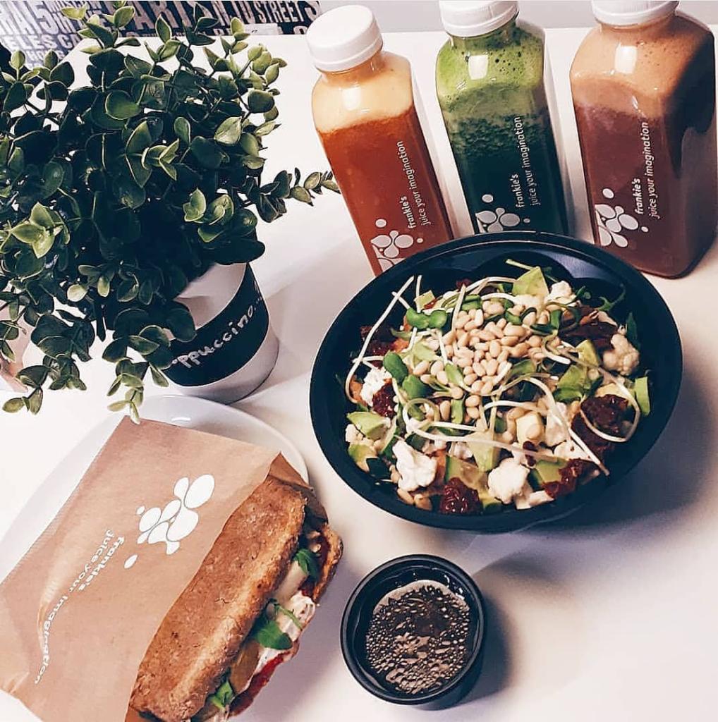 Kompozycja na białym tle: kanapka w papierowej torbie, sałatka z awokado i serem, trzy butelki kolorowych soków i roślina w doniczce z napisem 'Cappuccino'.