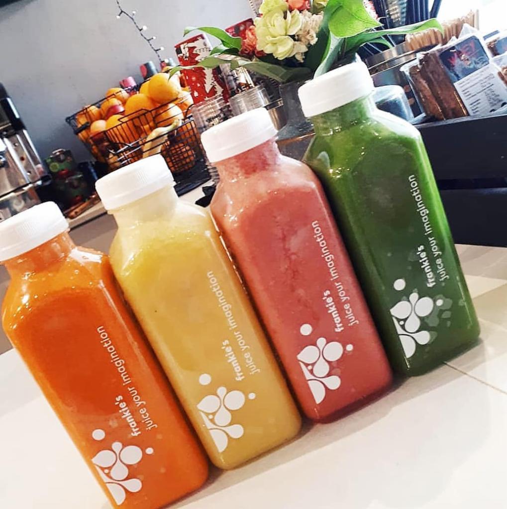 Cztery butelki świeżo wyciskanych soków o różnych kolorach (pomarańczowy, żółty, różowy, zielony) z logo 'Frankie's Juice your Imagination', ustawione ukośnie na jasnym blacie, w tle kosz...