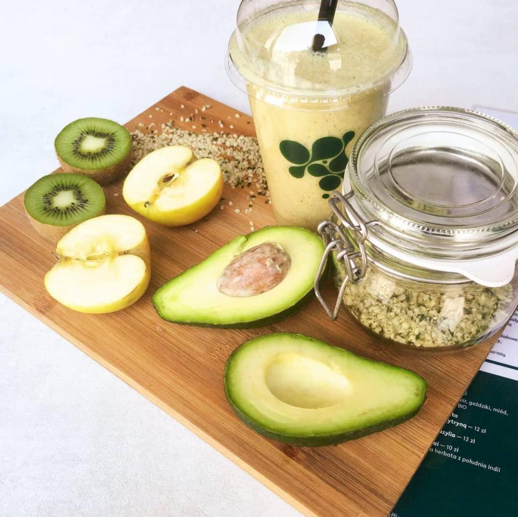 Kompozycja na drewnianej desce: przekrojone kiwi, jabłka, awokado, smoothie w plastikowym kubku z logo oraz słoik z nasionami, całość dopełnia karta menu z widocznymi cenami.