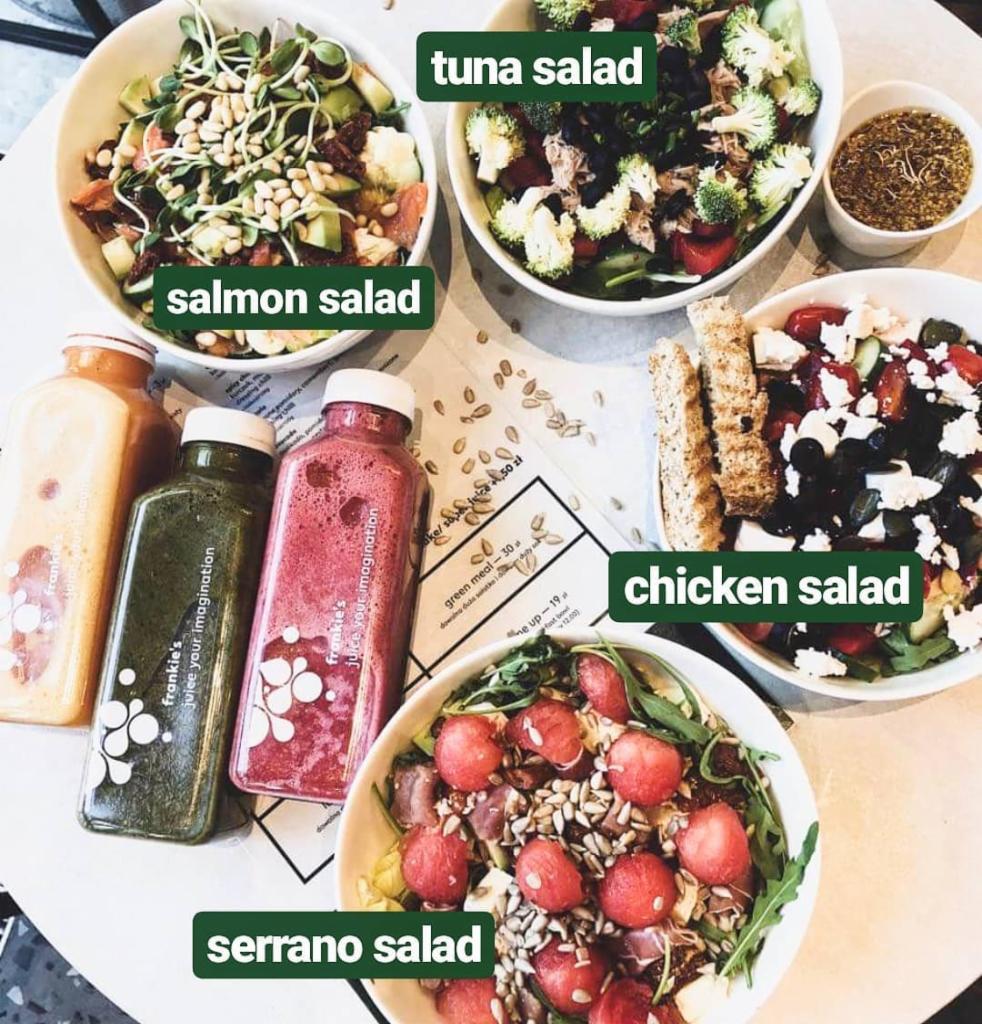 Różnorodne sałatki i soki Frankie's Juice w Poznaniu, w tym sałatka z łososiem, tuńczykiem, kurczakiem i serrano, ułożone na stole z menu, prezentowane w białych miskach i butelkach z logo.