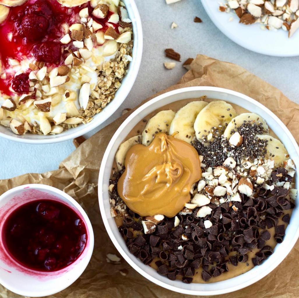 Dwa smoothie bowls na papierowym tle: jeden z jogurtem, granolą, migdałami i sosem wiśniowym, drugi z masłem orzechowym, bananem, nasionami chia, migdałami i kawałkami czekolady; dodatkowo miseczka...