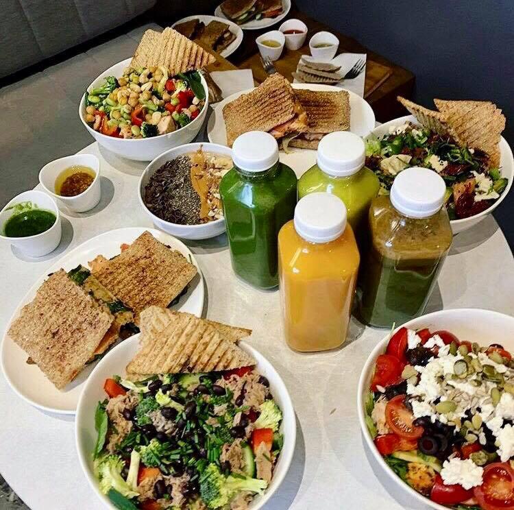 Kompozycja z różnorodnych sałatek, kanapek tostowych i świeżo wyciskanych soków w plastikowych butelkach, ułożonych na jasnym stole, z widocznym fragmentem szarej sofy w tle.