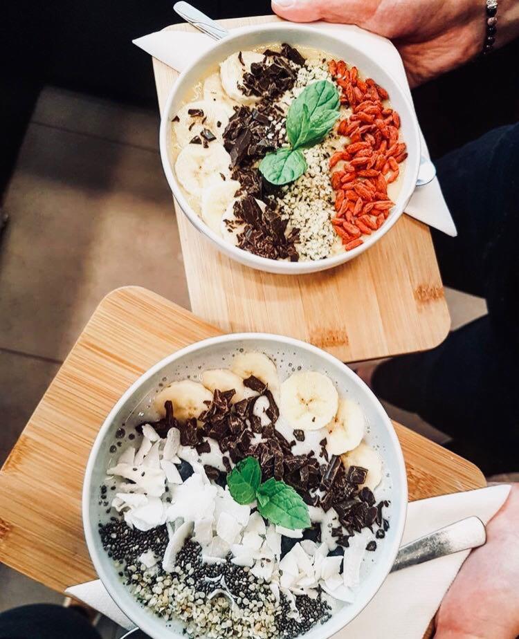 Dwie miski smoothie bowl z bananami, gorzką czekoladą, goji, wiórkami kokosowymi i miętą, podane na drewnianych deskach.