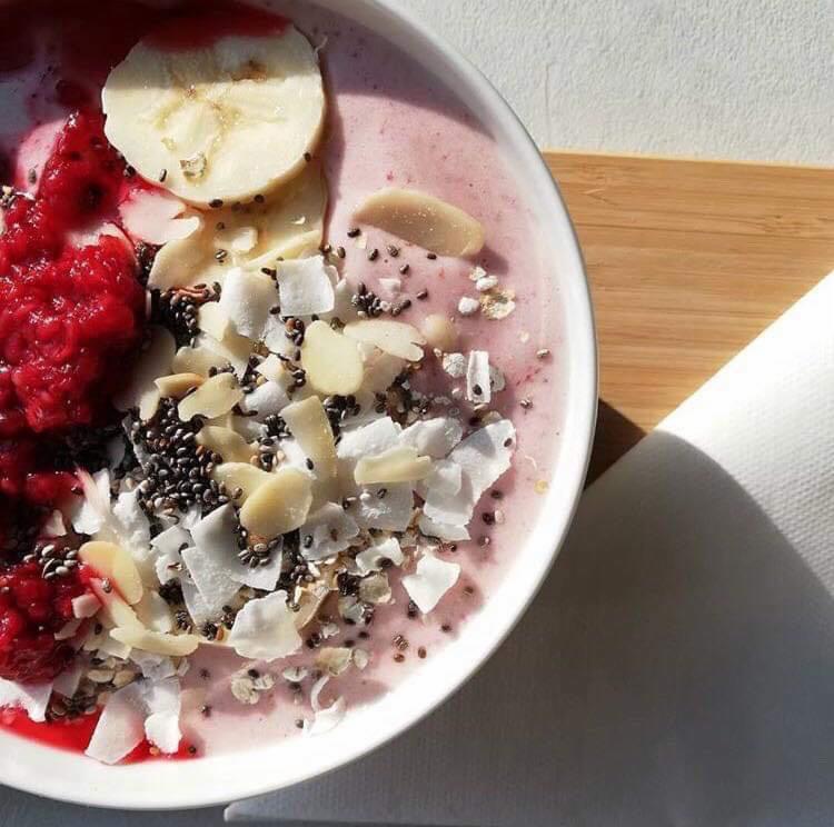 Różowy smoothie bowl z malinami, bananem, płatkami kokosowymi i nasionami chia, podany w białej misce na drewnianej desce, z widocznym cieniem serwetki.