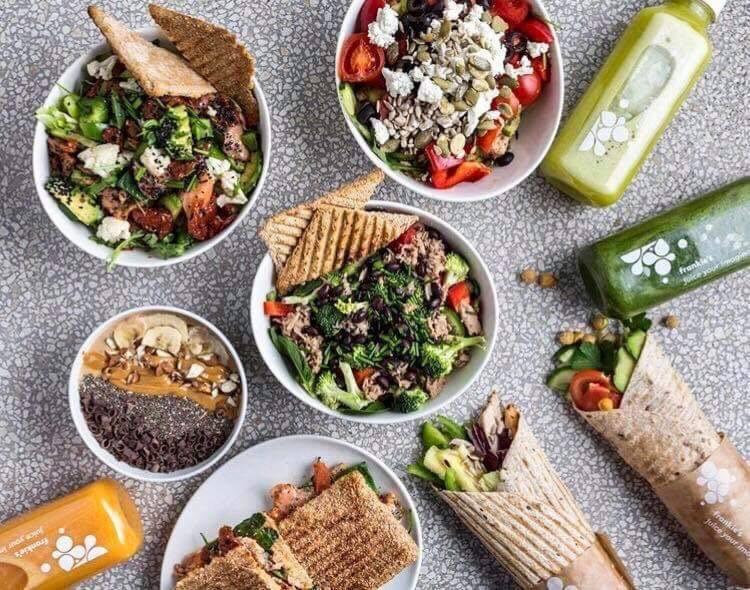 Kompozycja z różnorodnych dań: sałatki z warzywami i serem feta, bowl z bananami i masłem orzechowym, wrap z awokado i warzywami oraz kanapki na tle butelek ze świeżymi sokami.