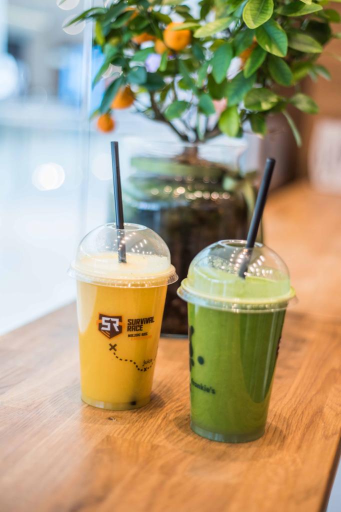 Dwa koktajle w plastikowych kubkach z czarnymi słomkami, jeden żółty z logo Survival Race, drugi zielony z logo Junkie's, na drewnianym blacie, w tle roślina w szklanym naczyniu.