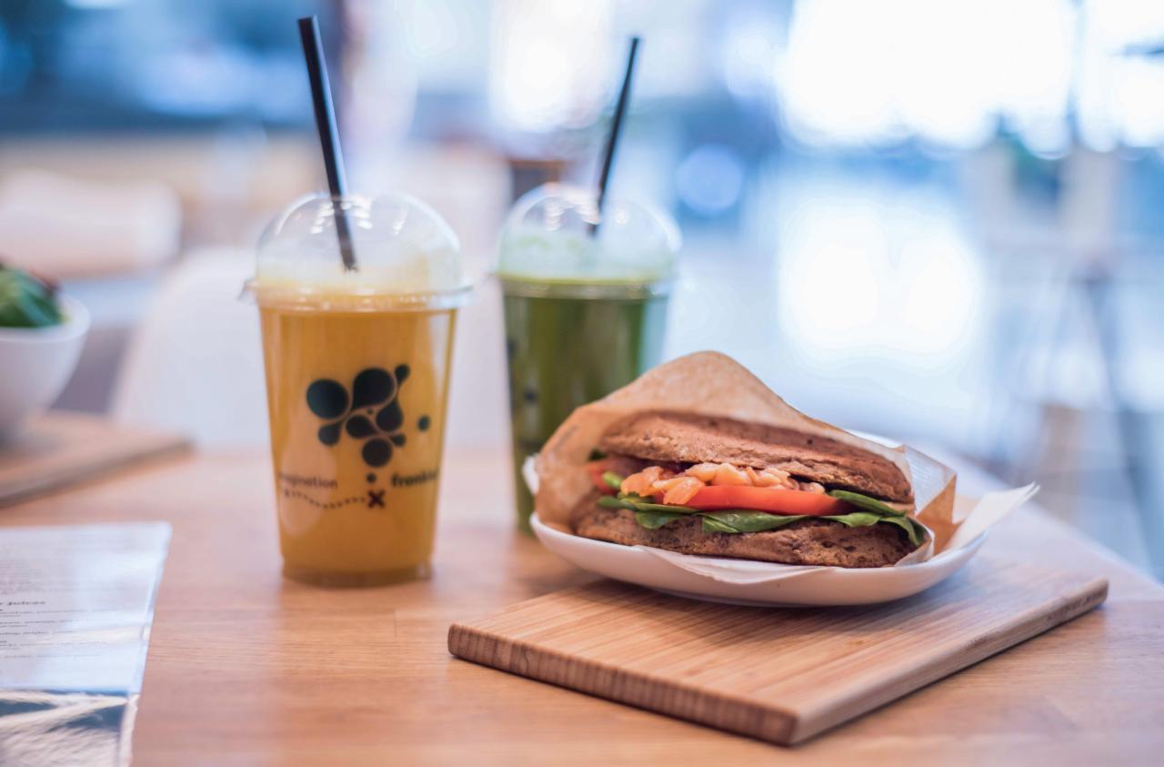 Kanapka z łososiem, pomidorem i szpinakiem na białym talerzyku, obok dwa napoje z czarnymi słomkami, jeden pomarańczowy z logo, drugi zielony, na drewnianym stole.