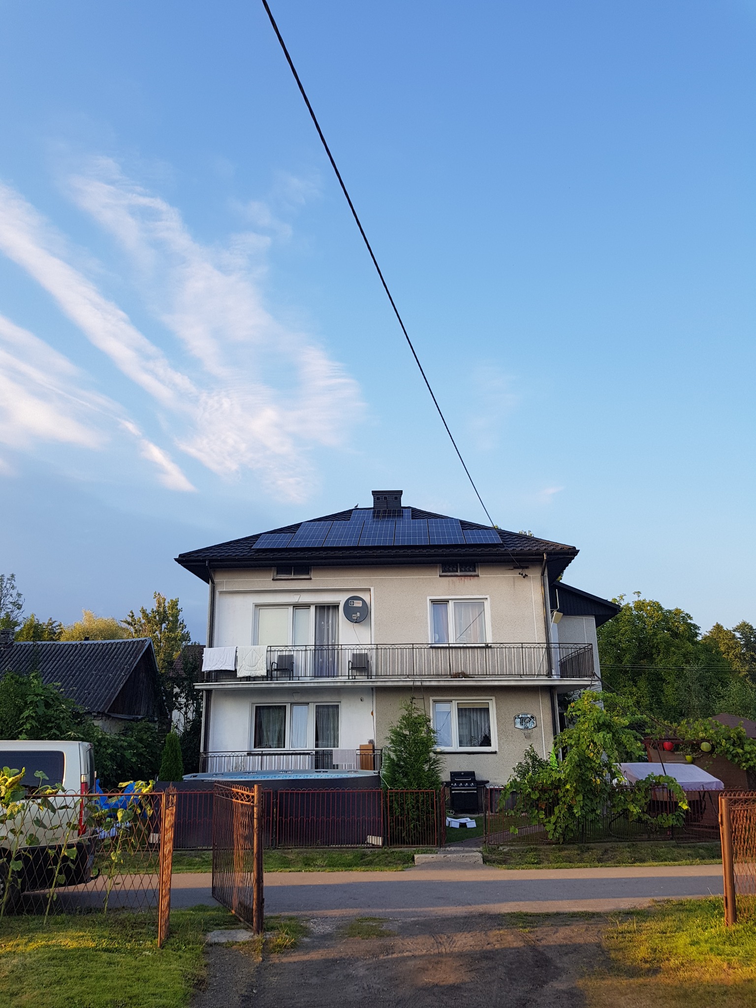 Dwupiętrowy dom z panelami słonecznymi na dachu, widoczny z poziomu ulicy przez otwartą, metalową bramę. Na balkonie suszą się białe ręczniki. W tle błękitne niebo z delikatnymi obłokami.