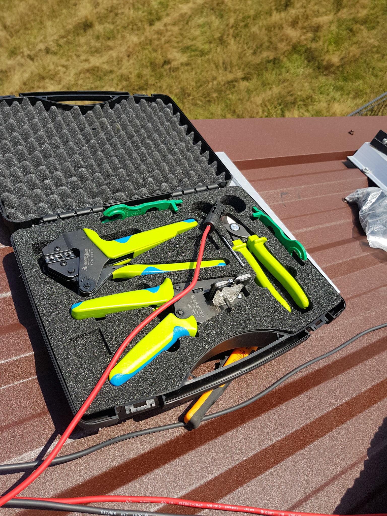 Otwarta czarna walizka z narzędziami do instalacji fotowoltaicznych, w tym zaciskarki z zielono-żółtymi uchwytami i przewody, leżąca na brązowym dachu z blachy trapezowej w słoneczny dzień.