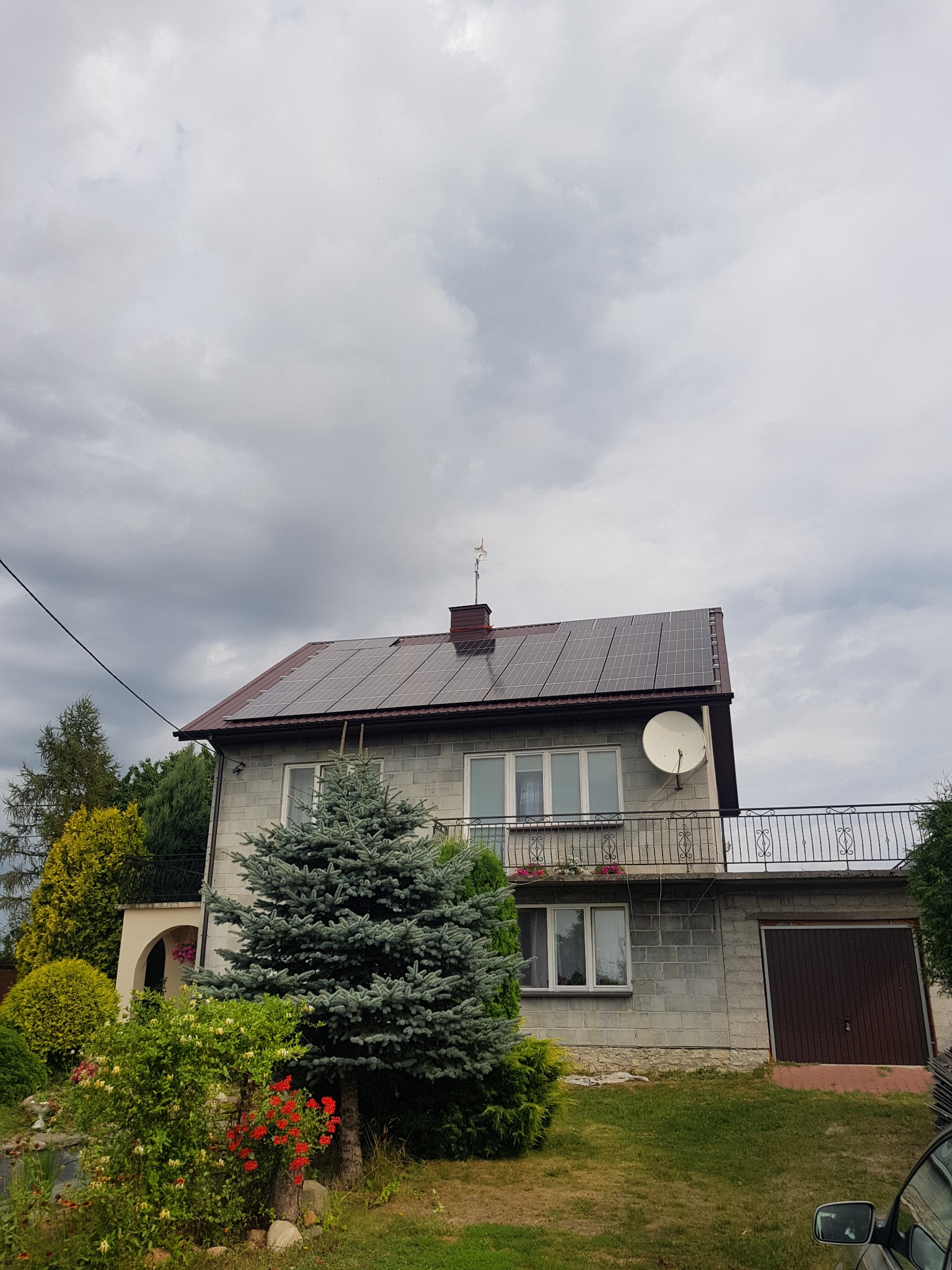 Dwupiętrowy dom z szarego pustaka z brązowymi panelami słonecznymi na dachu i anteną satelitarną na balkonie, otoczony zielenią i drzewami iglastymi