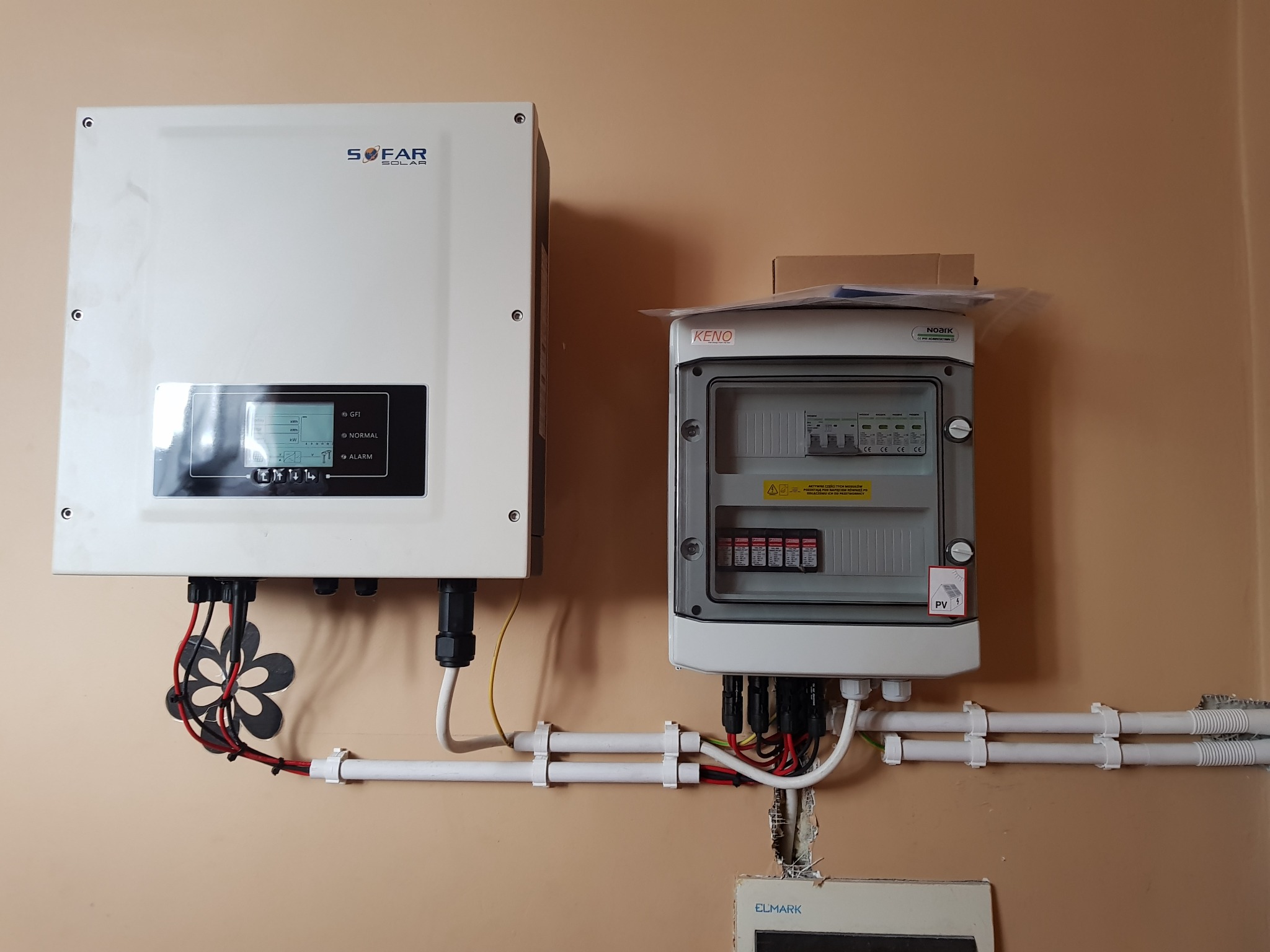 Biały falownik Sofar Solar z wyświetlaczem i rozdzielnica elektryczna Keno zamontowane na beżowej ścianie, widoczne przewody w peszlach i ozdobny element maskujący kable w kształcie kwiatka.