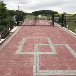 Czerwona nawierzchnia z szarej kostki betonowej ułożona we wzór przed czarną, automatyczną bramą wjazdową na posesję z widokiem na pagórkowaty krajobraz.