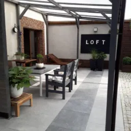 Zadaszony taras z szarą kostką, drewnianą pergolą, meblami ogrodowymi i dekoracjami w stylu loft.