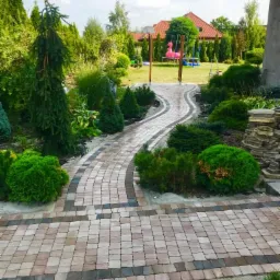 Wzorzysta ścieżka z kostki brukowej w ogrodzie, prowadząca do huśtawki z różowym flamingiem w tle.