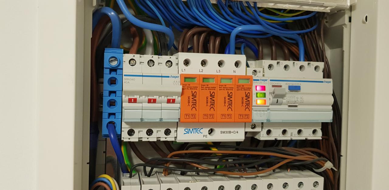 Rozdzielnica elektryczna z zainstalowanymi zabezpieczeniami przepięciowymi marki Simtec, widoczne przewody w różnych kolorach podłączone do listew zaciskowych i wyłączników, zamontowana w białej...