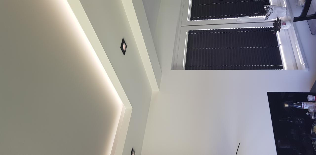 Nowoczesne oświetlenie sufitowe z wbudowanymi punktami świetlnymi i liniowym podświetleniem LED, widok w kierunku okna z żaluzjami plisowanymi.