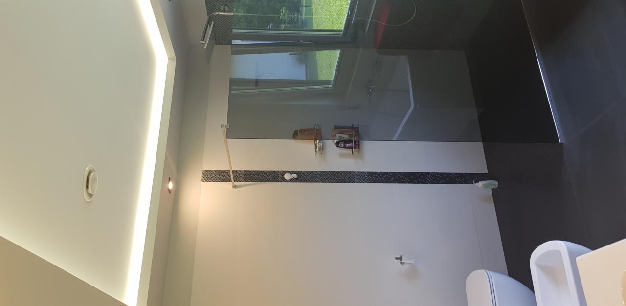 Nowoczesna łazienka z przeszklonym prysznicem typu walk-in, wykończona białymi płytkami z poziomym pasem dekoracyjnej mozaiki i czarnymi płytkami na podłodze, widok na zewnątrz przez okno.