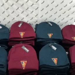 Rządki granatowych i bordowych czapek zimowych z wyhaftowanym herbem Pogoń Szczecin, ułożone na papierze z napisem 'Naszyj To'.