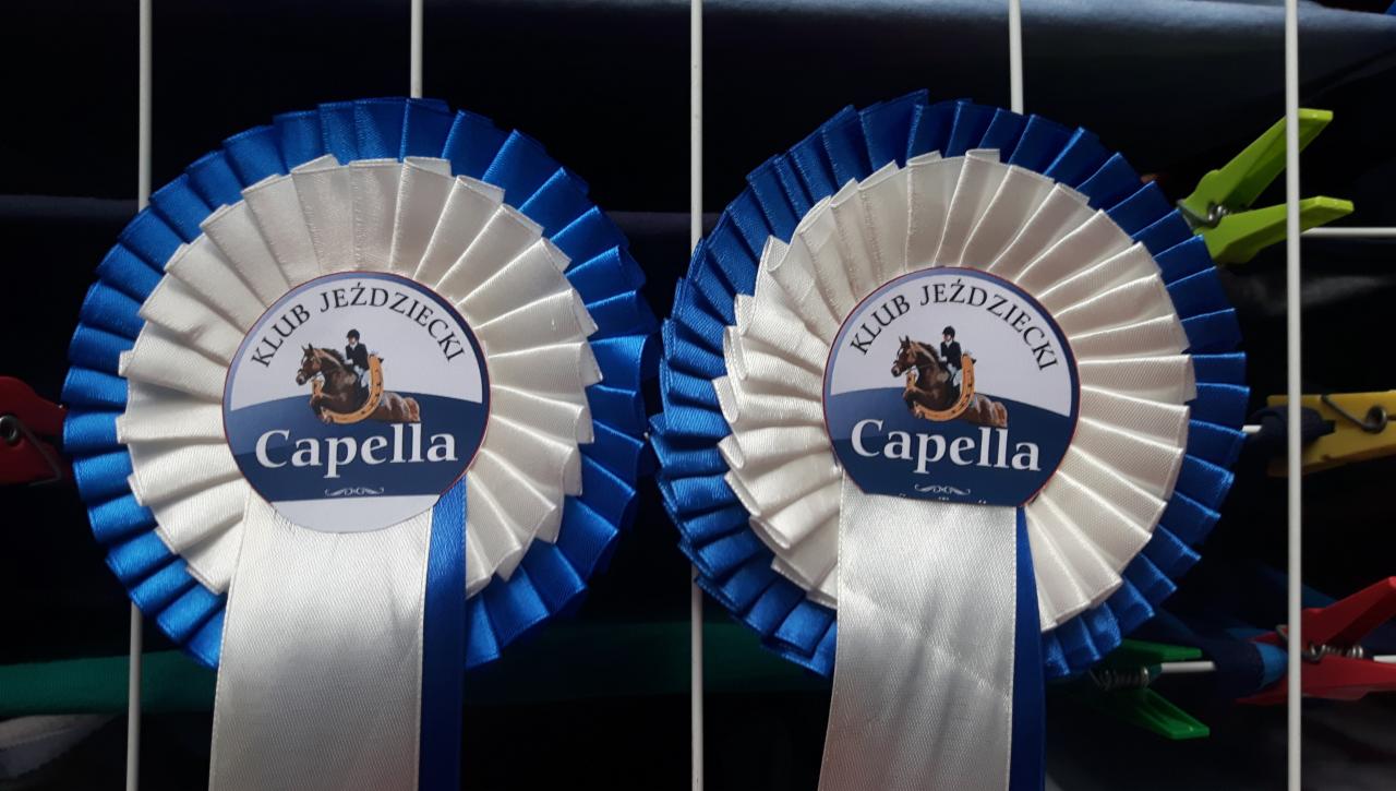Dwa identyczne rozetowe medale z napisem 'Klub Jeździecki Capella' w kolorach białym i niebieskim, prawdopodobnie zdobyte w Wąsowie, przypięte klamerkami do suszarki na pranie.