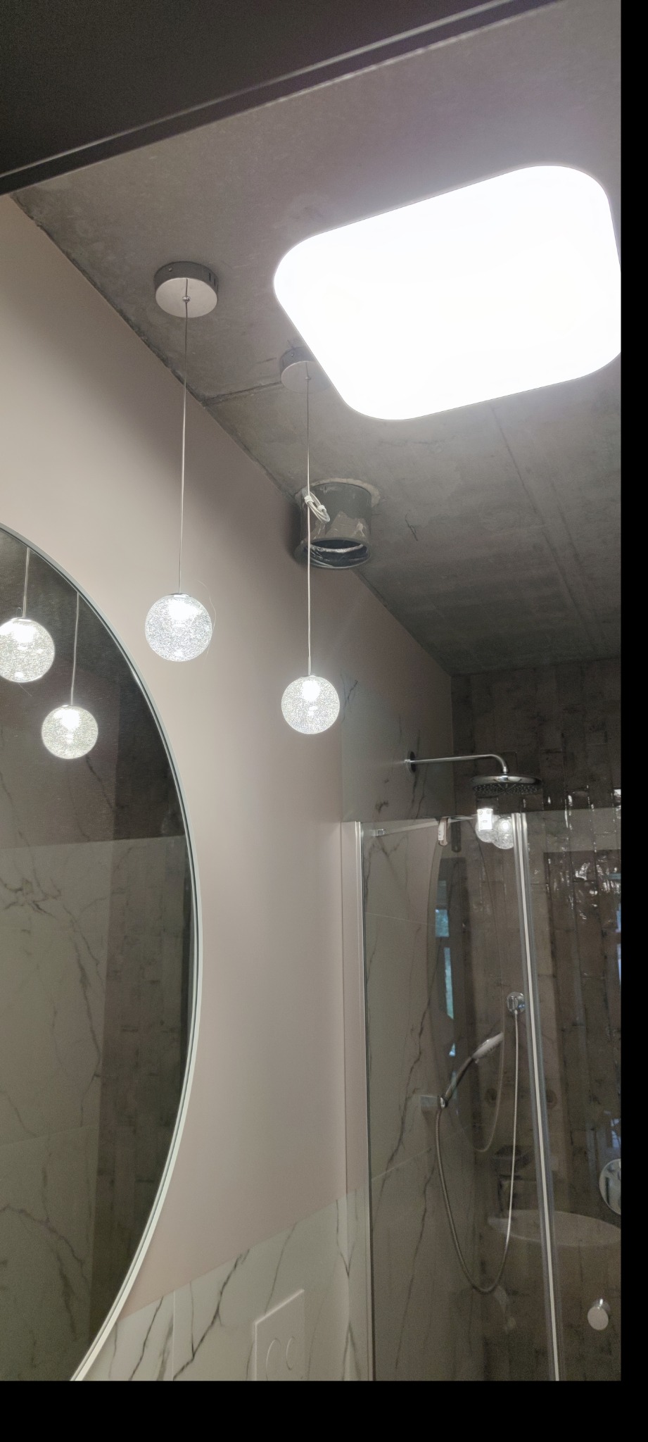 Nowoczesna łazienka z betonowym sufitem, okrągłym lustrem i wiszącymi lampami kulami, widoczna instalacja elektryczna przy suficie.