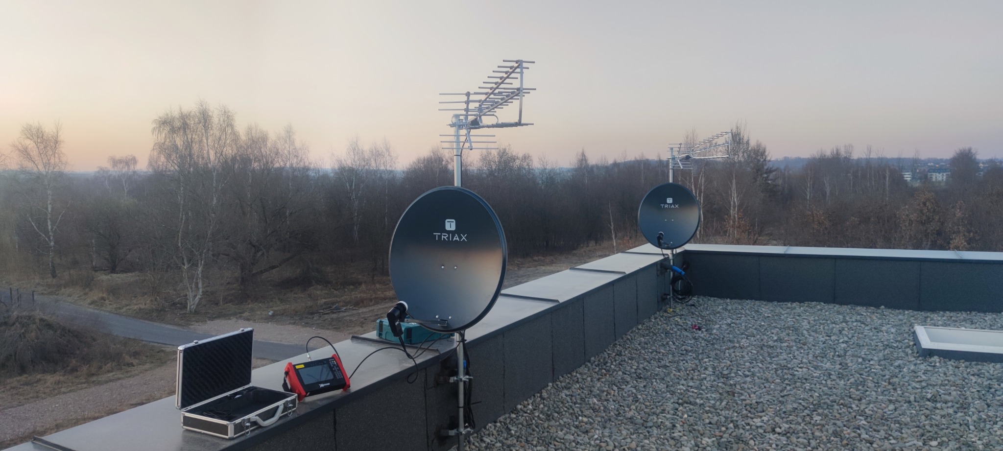 Dwie anteny satelitarne Triax i antena naziemna na dachu budynku, widoczne narzędzia pomiarowe w otwartym kufrze, tło stanowią drzewa i zachodzące słońce.