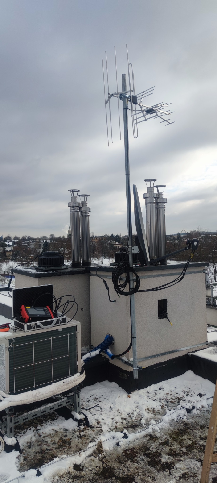 Antena telewizyjna, antena satelitarna i kominy wentylacyjne na dachu budynku w zimowej scenerii, widoczny miernik elektryczny w walizce