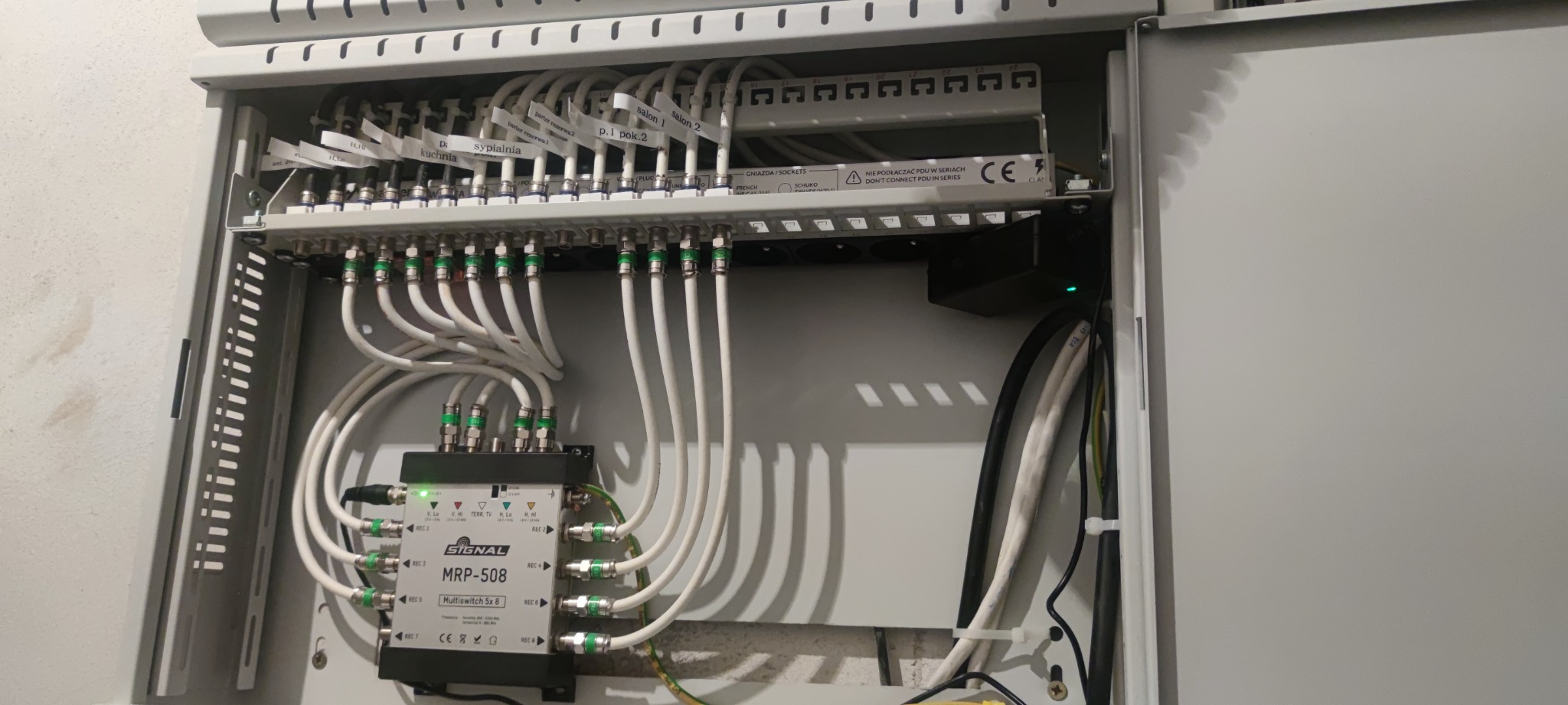Rozdzielnica z zainstalowanym multiswitchem Signal MRP-508, licznymi kablami koncentrycznymi z zielonymi złączkami oraz listwą zasilającą z gniazdami Schuko i French, opisana etykietami 'salon 1'...