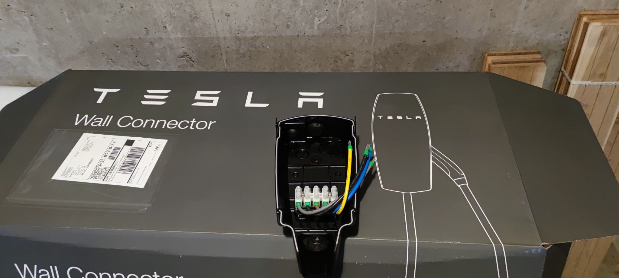 Otwarta skrzynka Tesla Wall Connector z widocznym wnętrzem i podłączonymi przewodami elektrycznymi w kolorach żółtym, niebieskim i zielonym, na tle betonowej ściany i drewnianych elementów.