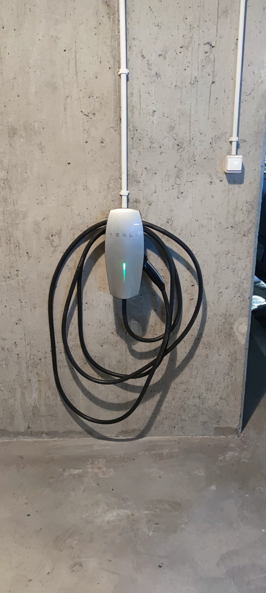 Biała ładowarka Tesla zamontowana na surowej, betonowej ścianie z widocznymi przewodami i zieloną diodą sygnalizującą gotowość do ładowania.