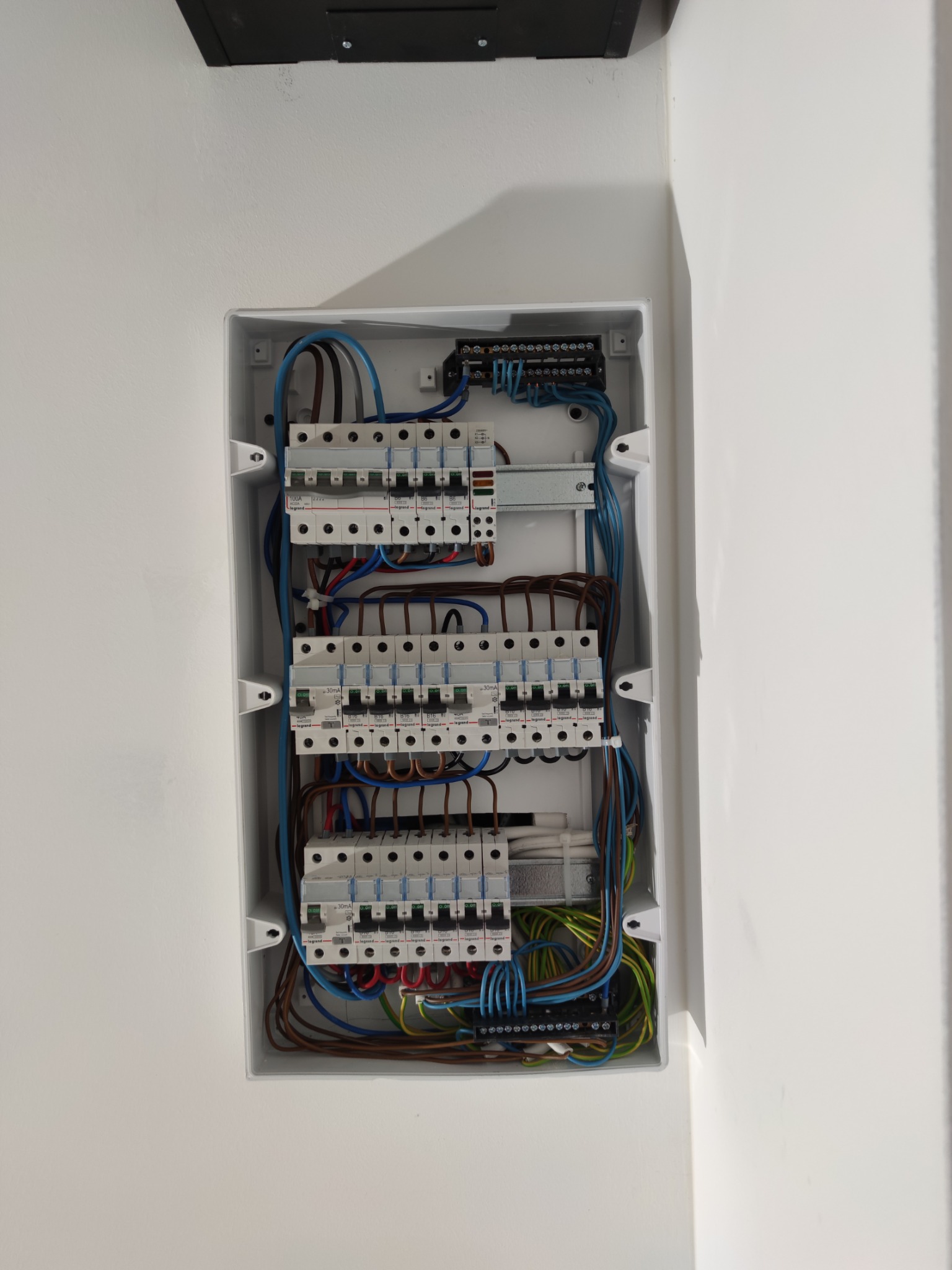 Otwarta skrzynka elektryczna z bezpiecznikami i przewodami w kolorach brązowym, niebieskim, zielonym i czarnym, zamontowana na białej ścianie.