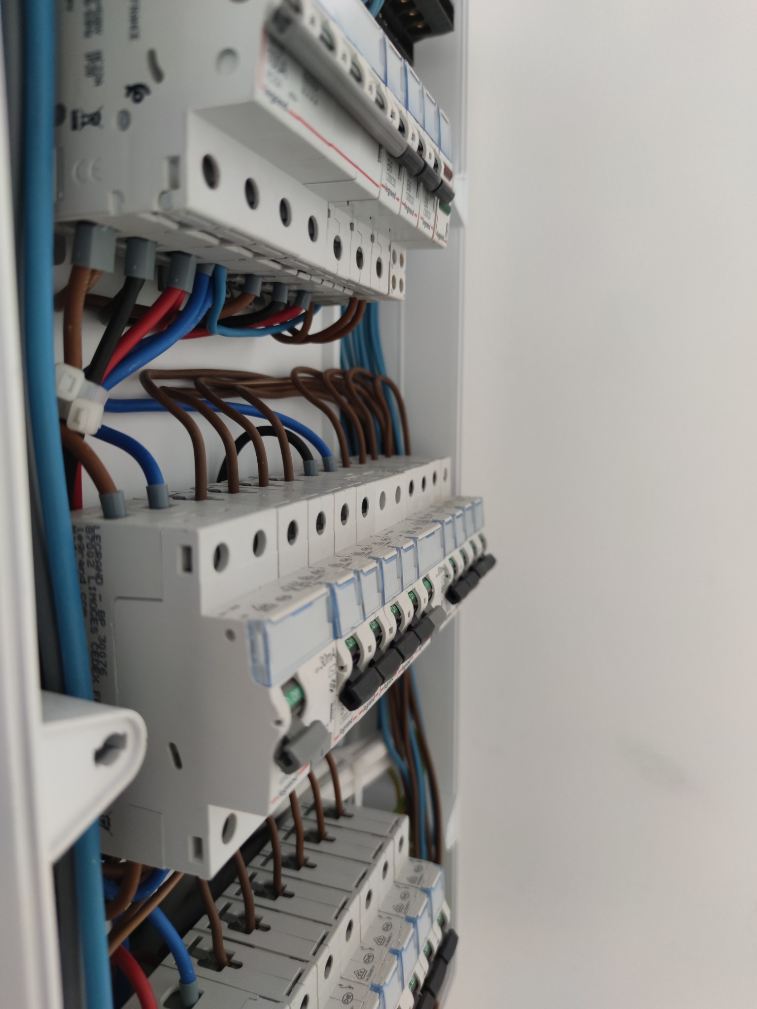 Szczegółowe ujęcie rozdzielnicy elektrycznej z widocznymi bezpiecznikami, przewodami w kolorach niebieskim, brązowym i czerwonym, oraz szyną DIN, zamontowanej w białej obudowie.