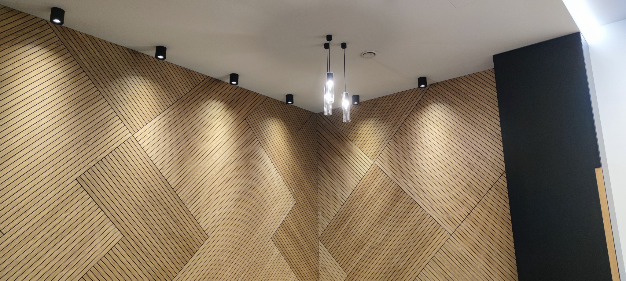 Wnętrze z drewnianą ścianą o geometrycznym wzorze, oświetloną przez czarne reflektory punktowe na suficie i wiszące lampy z przezroczystymi kloszami.