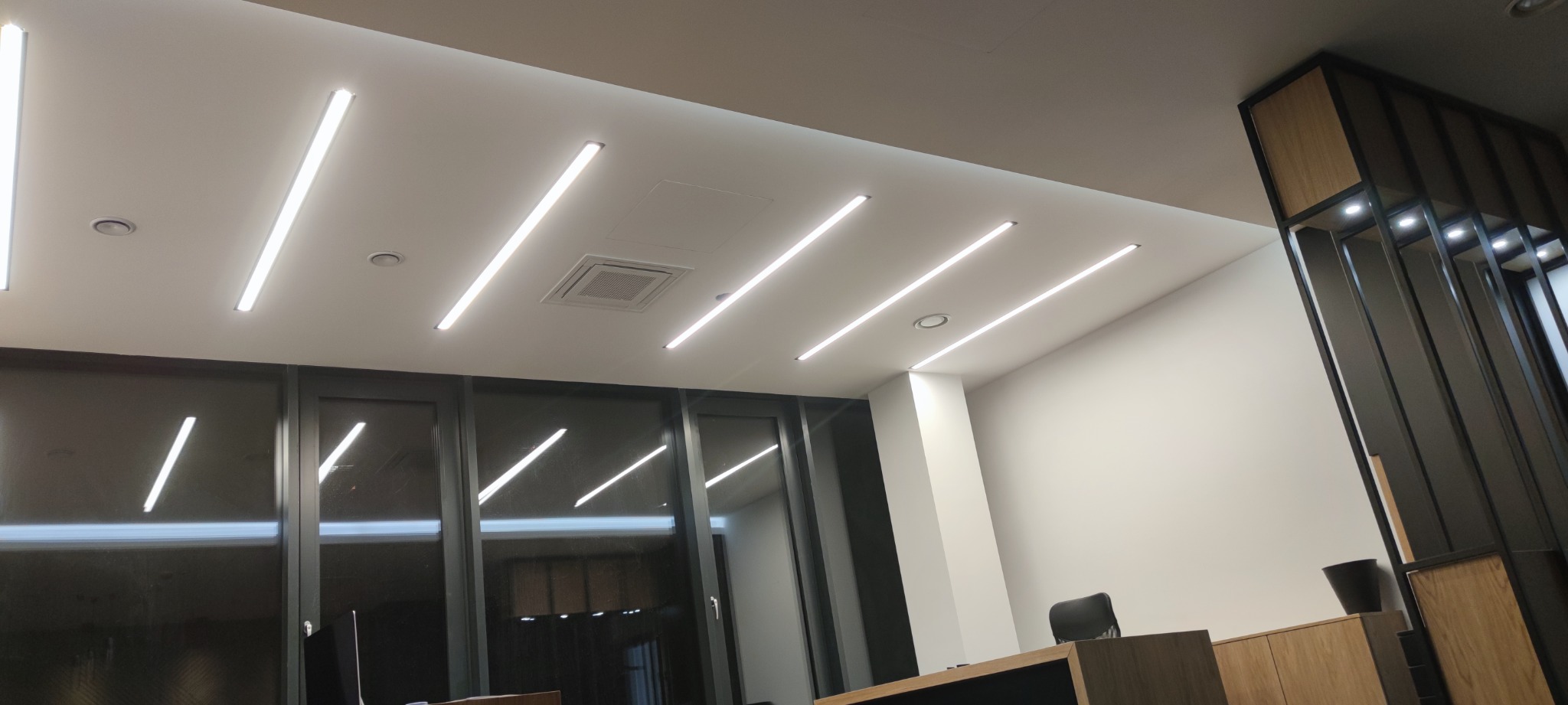 Nowoczesne biuro z równomiernie rozmieszczonymi liniowymi lampami LED wpuszczanymi w sufit, widoczna kratka wentylacyjna, duże okna i drewniane wykończenie ściany.