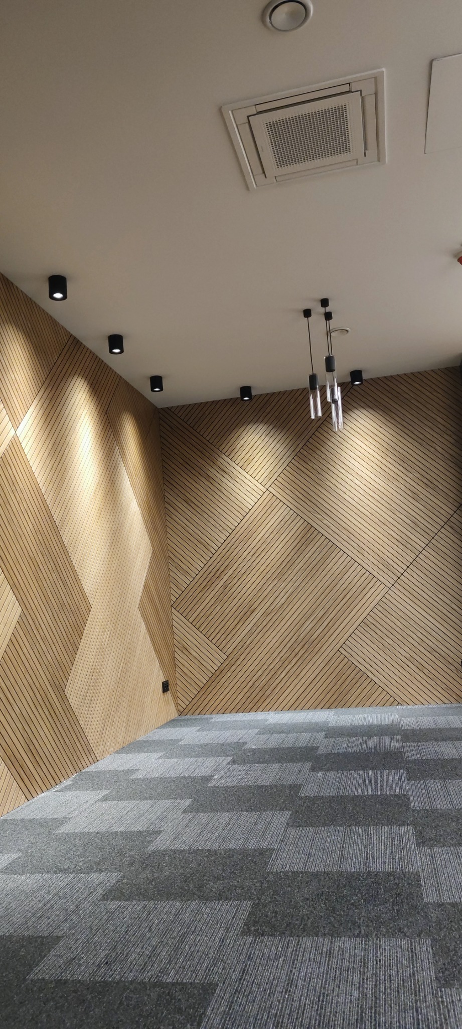 Wnętrze biurowe z drewnianą ścianą o geometrycznym wzorze, szarą wykładziną dywanową w kratkę i nowoczesnym oświetleniem sufitowym.