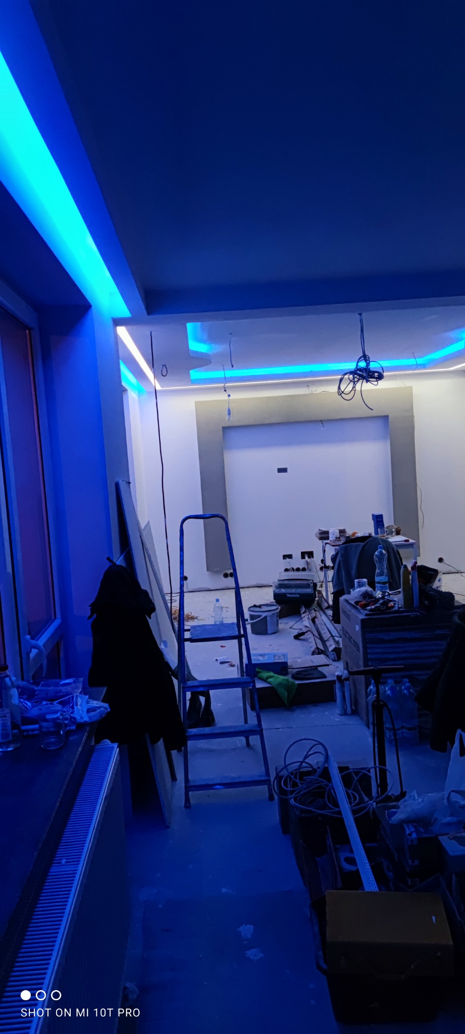 Niebieskie oświetlenie LED sufitowe w trakcie montażu w pokoju z drabiną i narzędziami, widoczne kable i niedokończone ściany.