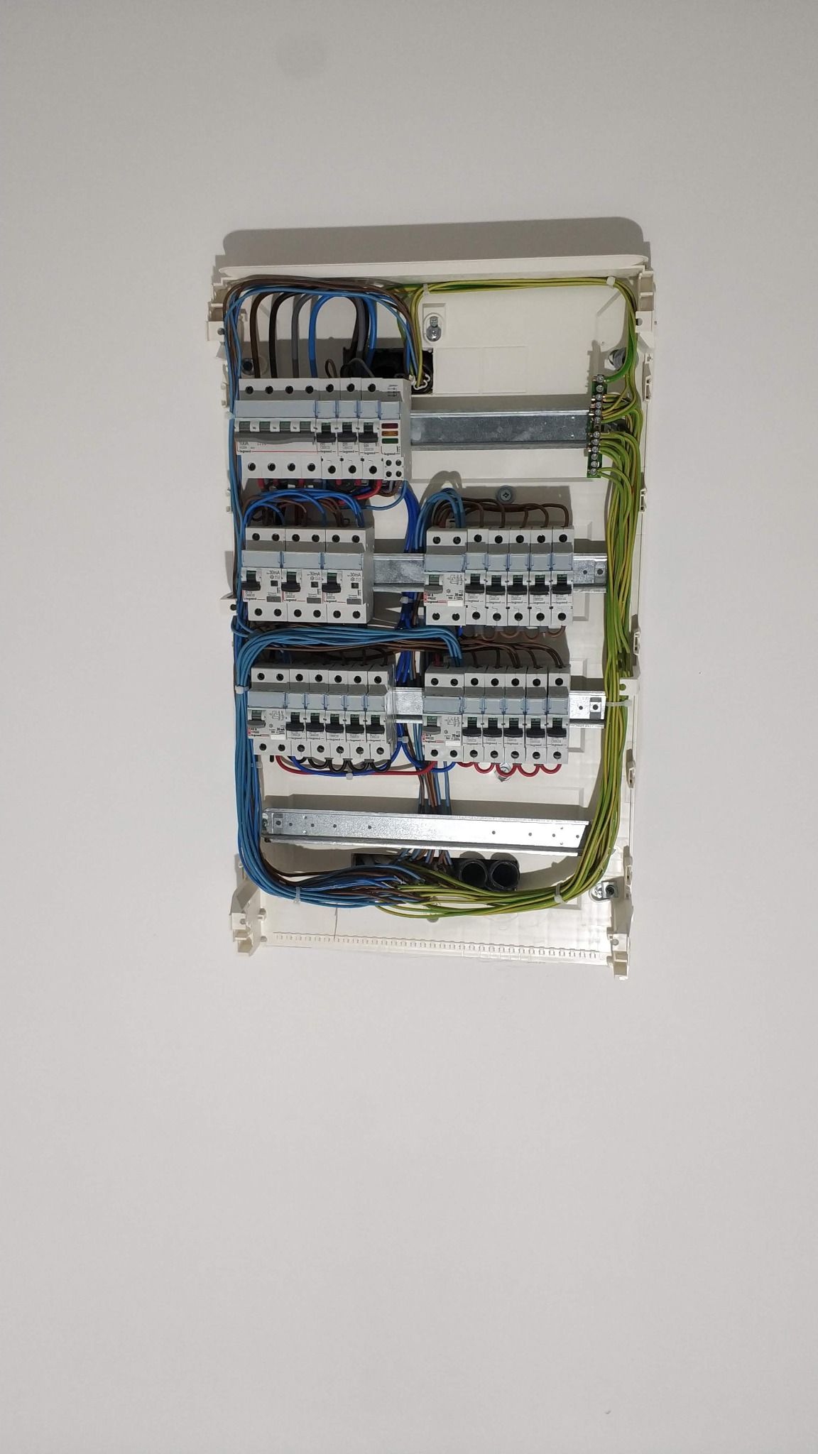 Otwarta skrzynka elektryczna z zainstalowanymi bezpiecznikami i podłączonymi przewodami w kolorach niebieskim, brązowym, zielono-żółtym i czerwonym, zamontowana na białej ścianie.