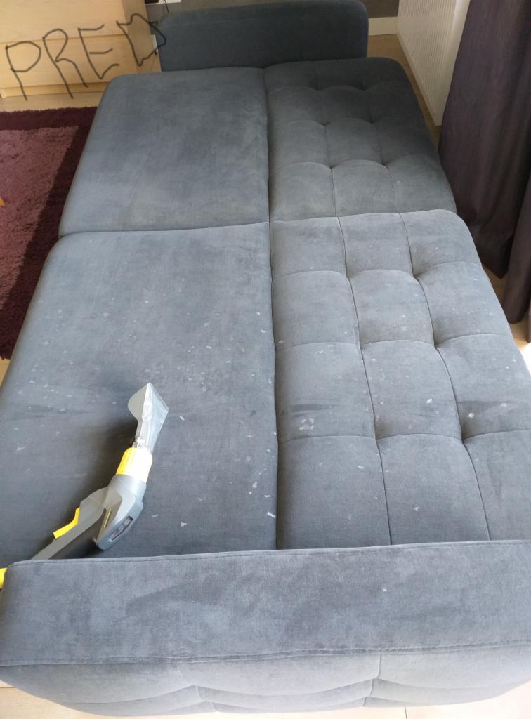 Szara, rozłożona sofa z widoczną różnicą po czyszczeniu: lewa strona z plamami, prawa po praniu, z dyszą odkurzacza piorącego na pierwszym planie.