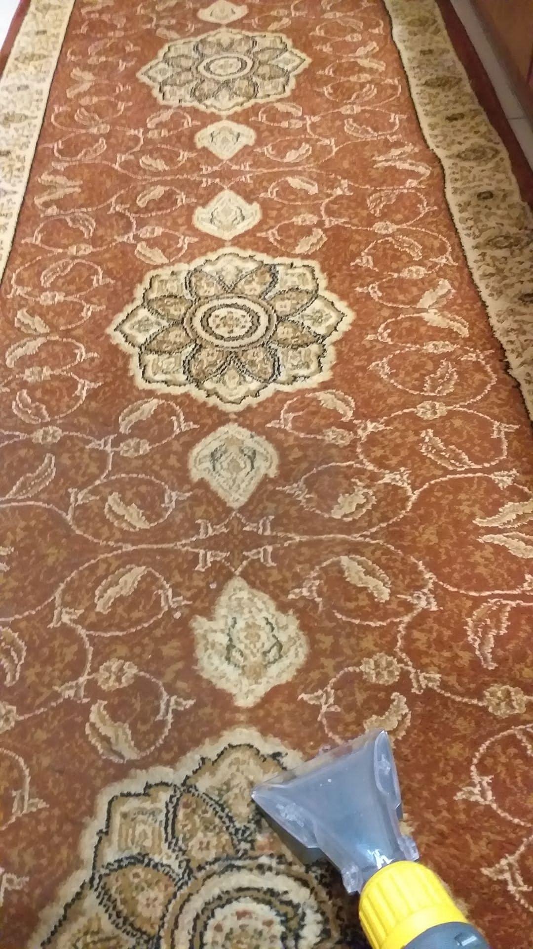 Fragment brązowego dywanu z orientalnym wzorem, czyszczonego przy użyciu końcówki odkurzacza piorącego z żółtą obudową.